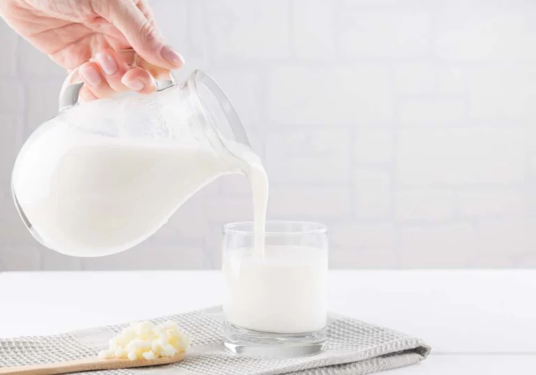 Milk poured from a glass jug into a glass, with kefir grains on a wooden spoon