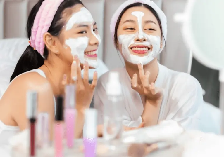 Two women in headbands smiling with white face masks applied, at a vanity with cosmetics